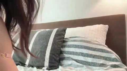 Video of a woman with long, dark hair, partially visible, lying on a bed with grey and white striped pillows. The room has a minimalist, modern decor with neutral tones.