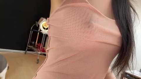 Video of a woman in a sheer, peach-colored, sleeveless top with a visible bra underneath. Her long, dark hair cascades over her shoulder. Background shows a black wall, a wooden floor, a desk, and a lamp.