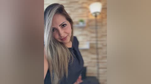 A video of a young woman with long, straight, blonde hair and a light complexion, wearing a black top, standing in a modern living room with a stone wall and a tall, white lamp.