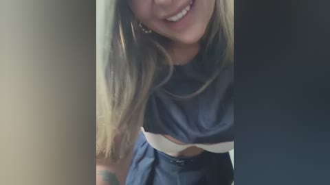 Media: Video of a young woman with light skin and straight, long blonde hair, smiling. She wears a dark blue crop top with a white midriff and a high-waisted skirt. The background is blurred, emphasizing her.