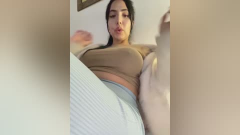A video of a Latina woman with medium skin tone, wearing a beige crop top and light grey high-waisted pants, sitting on a couch, blurred background.