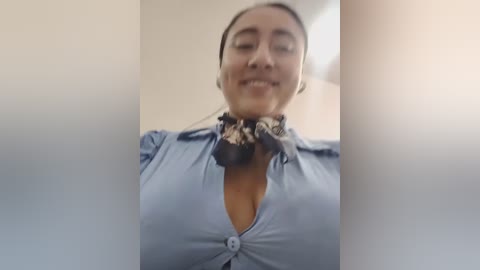 Media: Video of a smiling Latina woman with medium brown skin, wearing a blue button-up shirt, leopard-print scarf, and headphones, standing in a bright, indoor setting.