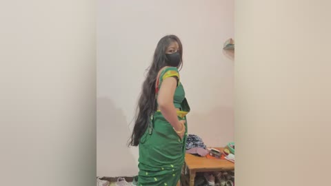 Video of a young woman with long black hair, wearing a green sari with yellow borders, black face mask, and black gloves, standing in a dimly lit, sparsely furnished room with a wooden table and scattered clothes.