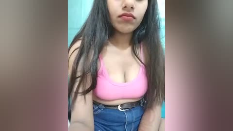 Video of a young woman with long, straight dark hair, wearing a pink sports bra and high-waisted blue jeans, standing in a bathroom with a teal-tiled background.