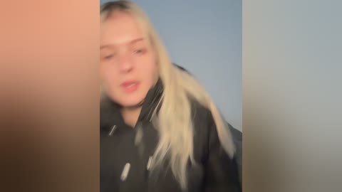 Video of a young, blonde woman with fair skin wearing a black leather jacket, blurred and out of focus, against a plain light blue background.