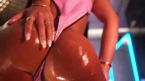 Video of a dark-skinned woman in a tight, pink bodysuit, highlighting her voluptuous, glistening buttocks. Her hand, adorned with a gold ring and manicured nails, rests on her hip. Background features blurred, neon blue lighting.