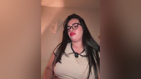 Video of a curvy, light-skinned woman with long black hair, wearing glasses, red lipstick, and a beige top, standing in a foggy, dimly-lit room.