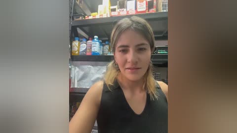 Video of a young woman with light skin, shoulder-length blonde hair, wearing a black sleeveless top, standing in a pharmacy aisle with shelves full of medicines and products.