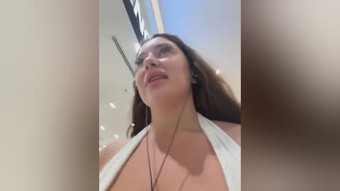 Video of a light-skinned woman with long brown hair wearing a white halter top, smiling upward. Background shows a modern, sunlit room with a skylight and a blurred man's face in the foreground.