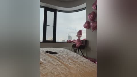 Video of a modern, minimalist bedroom with a large window showcasing a city skyline. A beige quilted bedspread covers the bed, and a pink teddy bear sits on a chair near the window. Pink balloons adorn the wall.