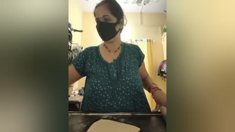 Media: A video of a woman wearing a black face mask, blue floral top, and bracelets, standing in a dimly lit kitchen with yellow walls.
