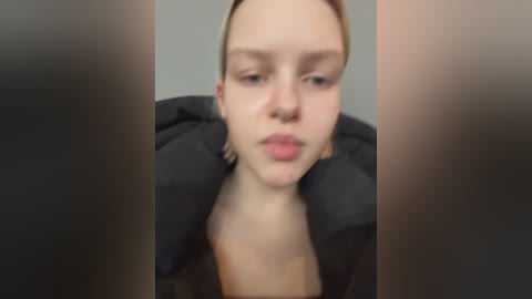 A video featuring a close-up of a young woman with fair skin, blonde hair, and a pouty expression, wearing a black coat, blurred by a focus on the foreground.