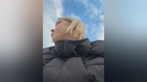 Video of a young blonde woman in a black puffy jacket, tilted head back, gazing at a partly cloudy blue sky, taken from a low angle.