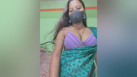 Video of a South Asian woman with medium skin tone, wearing a black face mask, purple lace bra, and green patterned sari, indoors with a bed and green wall in the background.