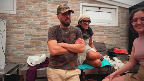 Video of a bearded man carrying a woman in a wheelchair, surrounded by a casual group in a room with stone-textured walls and clothes scattered on benches.