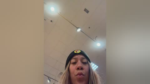 A video of a woman with a neutral expression wearing a black beanie with a yellow smiley face, standing in a dimly lit room with recessed ceiling lights.