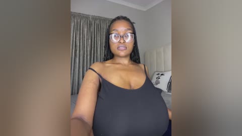 A video of a young Black woman with large breasts, wearing a black spaghetti-strap top and glasses, standing in a modern bedroom with beige walls and gray curtains.