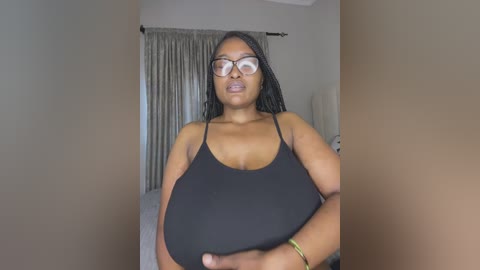 Video of a plus-sized Black woman with long braids, wearing glasses and a black tank top, standing in a beige room with curtains.