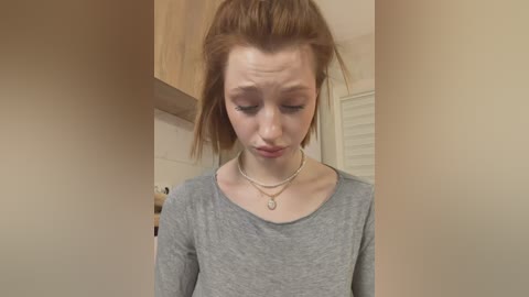 Video of a fair-skinned, red-haired woman with a high ponytail, wearing a gray long-sleeved shirt and a silver necklace, standing in a beige-walled room with wooden cabinets and white blinds.