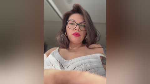 Video of a fair-skinned, curvy woman with dark hair, wearing glasses and a white off-shoulder knitted sweater, standing in a bright, minimalist room.