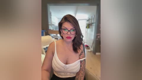 Video of a fair-skinned, dark-haired woman with glasses, wearing a white, sleeveless top, revealing tattoos on her right arm. She sits in a cluttered, modern kitchen with white cabinets and wooden countertops.