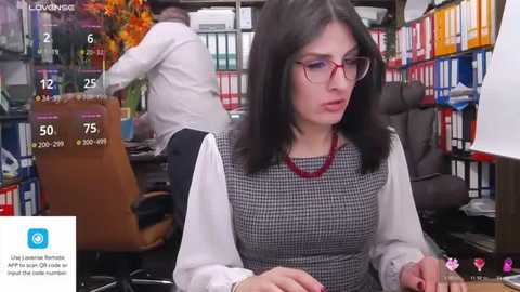 Media: A video of a serious woman with glasses and dark hair, dressed in a checkered blouse and red necklace, sitting in a cluttered office with shelves of books and a TV screen displaying weather and news.