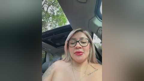 Video of a topless, plus-sized woman with light skin, straight blonde hair, and black-rimmed glasses, wearing a gold necklace, sitting in a car with a green tree background.