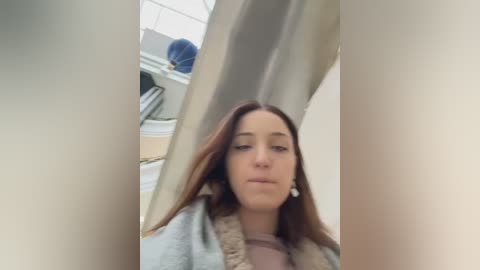 Video of a young woman with long brown hair, wearing a light jacket, looking through a large, light-colored curtain. Background shows a laundry room with white cabinets and laundry baskets.