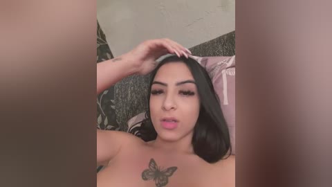 Video of a Latina woman with long black hair, pink lips, and a butterfly tattoo on her chest, sitting on a bed with floral-patterned sheets, adjusting her hair.