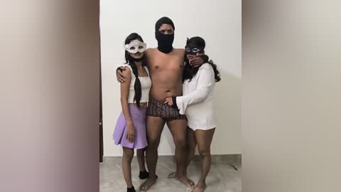 Video of a shirtless, muscular Black man wearing a black mask and boxer shorts, flanked by two women in white masks, one in a purple skirt, and the other in a white dress, standing in a plain room.