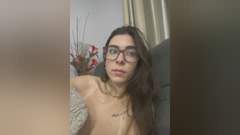 A video of a topless woman with glasses, long brown hair, and a tattoo, standing in a dimly lit room with beige curtains and a vase of red flowers.