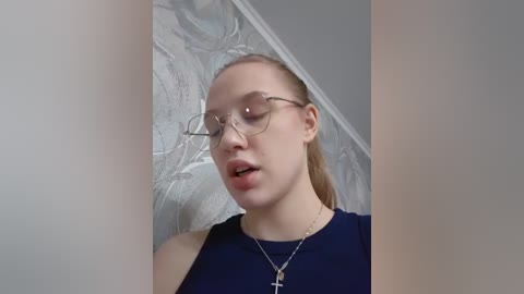 Media: Video of a young, fair-skinned woman with blonde hair, wearing large round glasses, a navy blue sleeveless top, and a silver cross necklace. Background features ornate, light-colored wallpaper with a floral design.