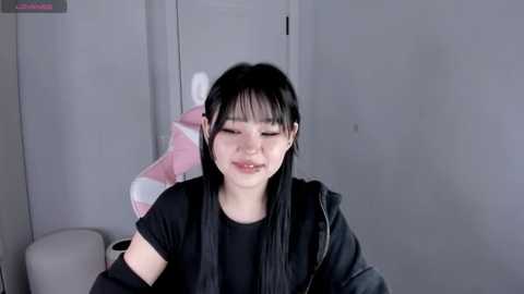Video of a young Asian woman with straight black hair and bangs, wearing a black jacket and shirt, smiling softly, in a minimalist gray room with a pink towel and tissue box in the background.