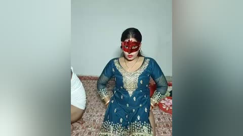 Media: A video of a woman in a dark blue, intricately embroidered Punjabi dress with red face mask, seated on a floral-patterned bed against a plain wall.