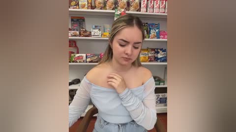 Video of a young woman with light skin and blonde hair, wearing a light blue off-shoulder top, standing in a brightly lit grocery store aisle filled with snacks.