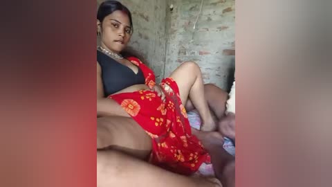 Media: Video of a young woman in a red floral dress, black bra, sitting on a bed, surrounded by brick walls.