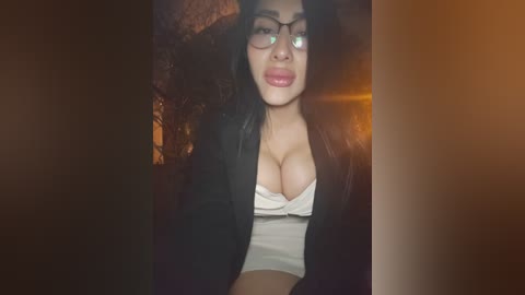 A video of an Asian woman with long black hair, wearing glasses, a black jacket, and a revealing white top, sitting in a dimly lit room with warm lighting.