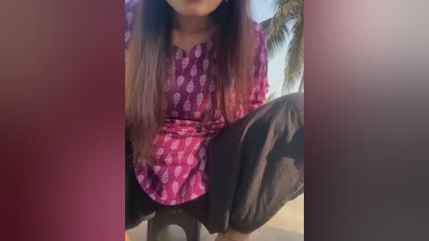 A video of a woman squatting, wearing a maroon top with white floral patterns, black pants, and long dark hair. The background shows a palm tree and clear sky.