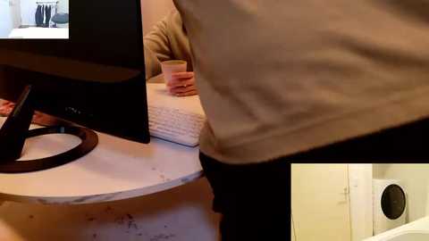 A video depicts a person in a beige sweater, standing in a kitchen, facing a computer monitor on a white marble countertop. The background shows a pink cup and a blurred person seated.