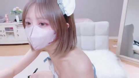 Media: Video of an East Asian woman with light skin, wearing a white face mask and a white and blue checkered headband, in a modern bedroom with light wooden floors and a white bed.