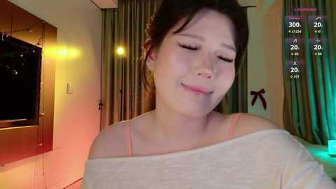 Media: A video of a smiling Asian woman with fair skin and dark hair, wearing a white off-shoulder top, standing indoors. Background includes a TV, curtains, and a digital overlay showing time and date.