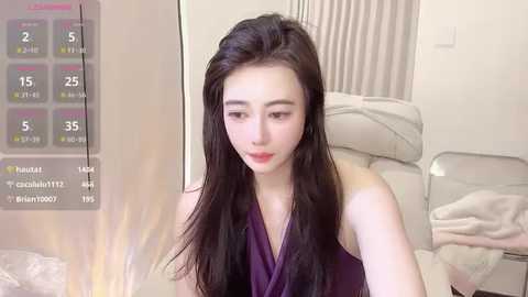 Media: Video of a young Asian woman with long black hair, wearing a purple sleeveless dress, sitting on a bed in a modern, minimalistic room with a digital temperature display.