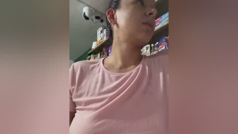 Video of a pregnant woman with medium-dark skin and straight, dark hair tied back. She wears a light pink ribbed tank top. Background shows a messy room with a ceiling fan, shelves of colorful items, and a white wall.