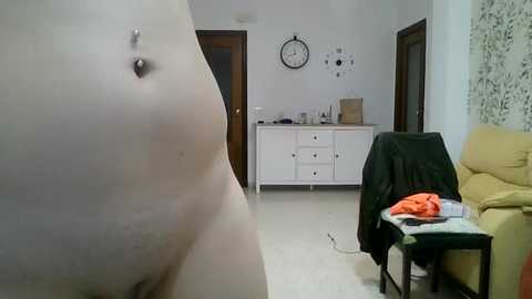 A video showing a close-up of a woman's bare abdomen with a belly button piercing, standing in a cluttered living room with a white dresser, clock, and beige sofa.