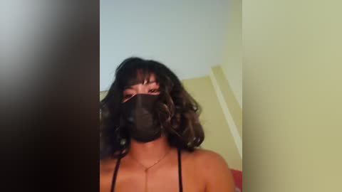 Media: Video of a woman with shoulder-length curly black hair, wearing a black face mask and black halter top, standing in a dimly lit room with beige walls and a red couch.