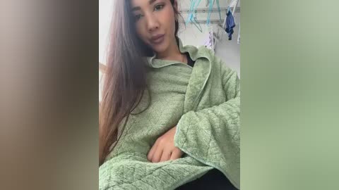 Video of a young woman with long, straight brown hair, wearing a light green, textured robe, sitting in a bathroom with hanging clothes and towels.
