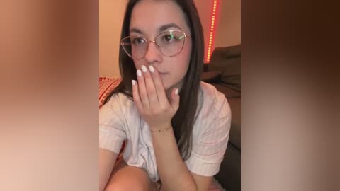 Video of a young woman with fair skin, long dark hair, and glasses, covering her mouth with her hand. She wears a light-colored shirt and sits on a bed with a striped pillow in a cozy, dimly-lit room with red LED lights.