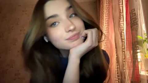 Video of a young woman with long brown hair, fair skin, and blue eyes, resting her chin on her hand. She's indoors near a window with red and gold curtains.
