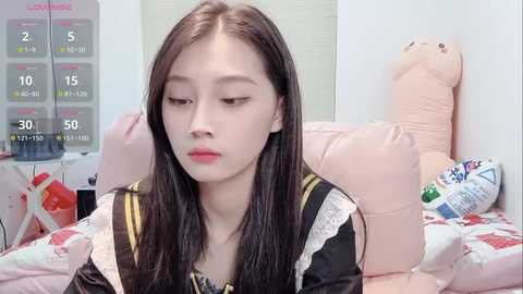 Video of a young Asian woman with long, straight black hair and fair skin, dressed in a black and white sailor uniform, sitting on a bed surrounded by plush toys, including a large pink bear.