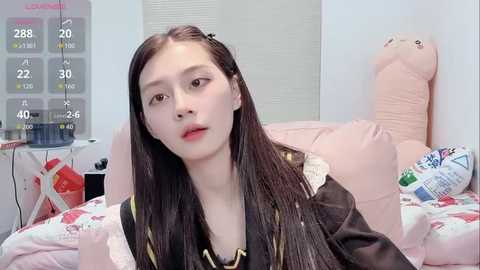 Video of a young Asian woman with long black hair, wearing a black jacket, sitting on a bed with a pink pillow and a plush toy. The background includes a calendar and a window.